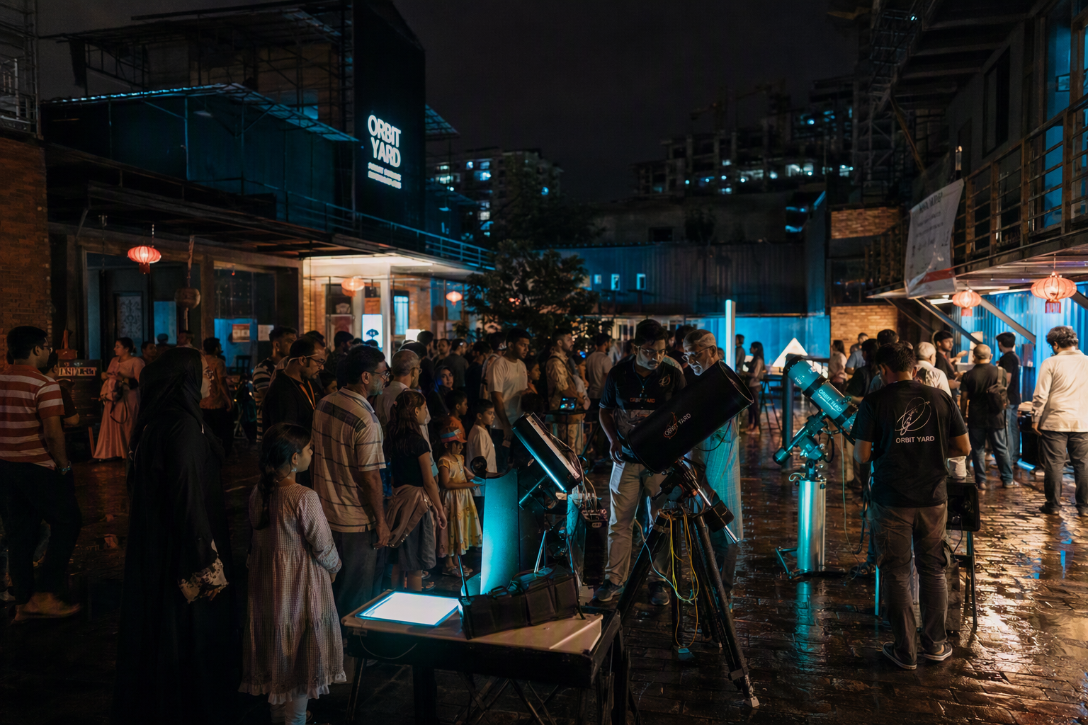 A lively evening crowd gathered around glowing science demonstrations in Orbit Yard's open courtyard under a deep blue sky